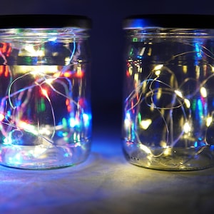 May include: Two clear glass jars with lids filled with white string lights. The jar on the left has multi-colored lights, while the jar on the right has white lights.