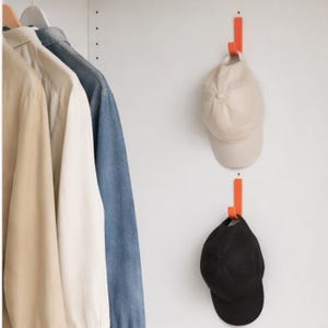 Puede incluir: Dos ganchos metálicos naranjas montados en una pared blanca, cada uno sosteniendo una gorra de béisbol. Una gorra es beige, la otra es negra. Ropa colgada en una percha a la izquierda.