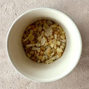 May include: A white ceramic bowl filled with light amber-colored, irregularly shaped resin pieces. The bowl is centered against a neutral, textured background. The resin pieces have a translucent quality.