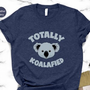 A navy blue t-shirt with a graphic of a koala and the text "Totally Koalified" printed on it, along with a pair of glasses and a book.