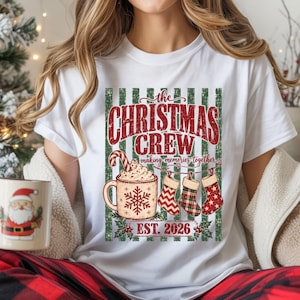 May include: A white t-shirt featuring a graphic with the text "the CHRISTMAS CREW making memories together EST. 2026". The design includes a mug with a snowflake and candy cane, three hanging stockings, and a green and white striped background.