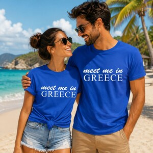 A man and a woman standing together on a beach, both wearing matching blue t-shirts with the text "Meet me in Greece".