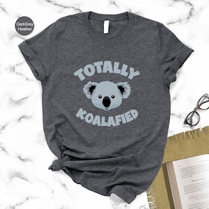 A gray t-shirt with a graphic of a koala and the text "Totally Koalified" printed on it, along with a pair of glasses and a book on a marble surface.