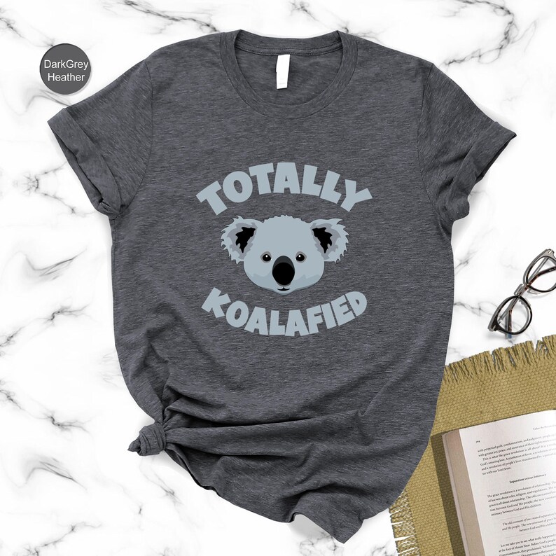 A gray t-shirt with a graphic of a koala and the text "Totally Koalified" printed on it, along with a pair of glasses and a book on a marble surface.