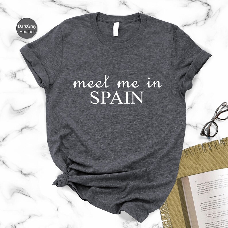 a gray t-shirt with the text "Meet me in Spain" printed on it, along with a pair of glasses and a book on a marble surface.