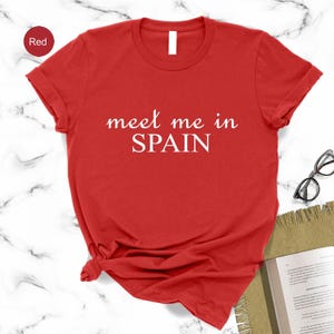 A red t-shirt with the text "Meet me in Spain" printed on it, along with a pair of glasses and a notebook on a marble surface.