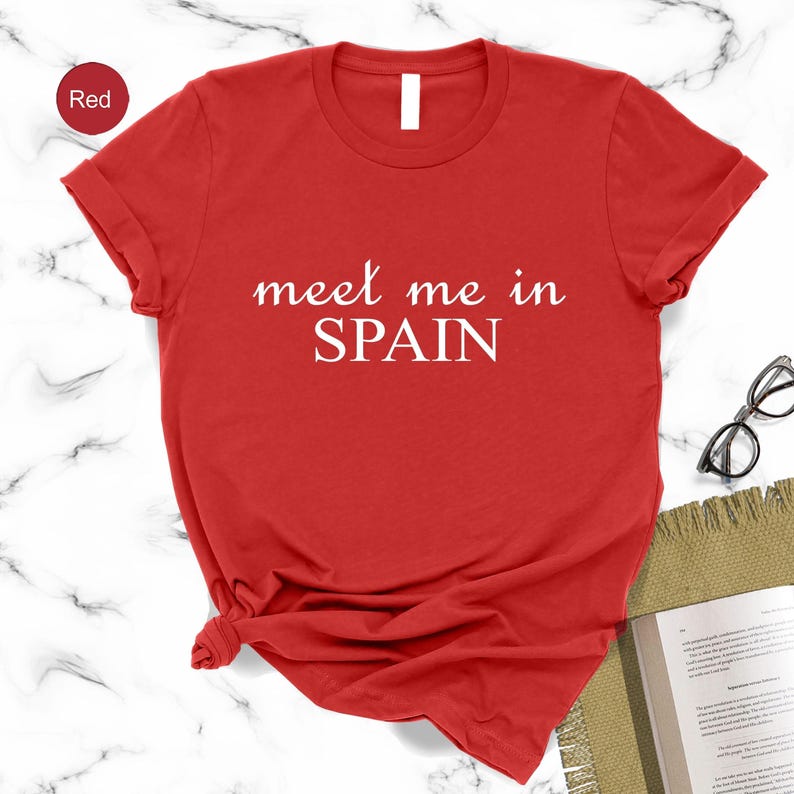 A red t-shirt with the text "Meet me in Spain" printed on it, along with a pair of glasses and a notebook on a marble surface.