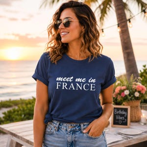 A woman wearing a blue t-shirt with the text "Meet me in FRANCE" stands near a table with flowers and a sign that says "Birthday Vibes".
