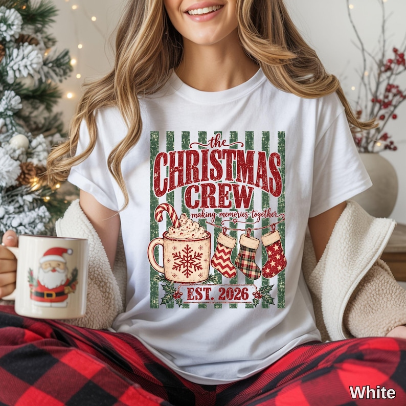 May include: A white t-shirt featuring a graphic with the text "the CHRISTMAS CREW making memories together EST. 2026". The design includes a mug with a snowflake and candy cane, three hanging stockings, and a green and white striped background.