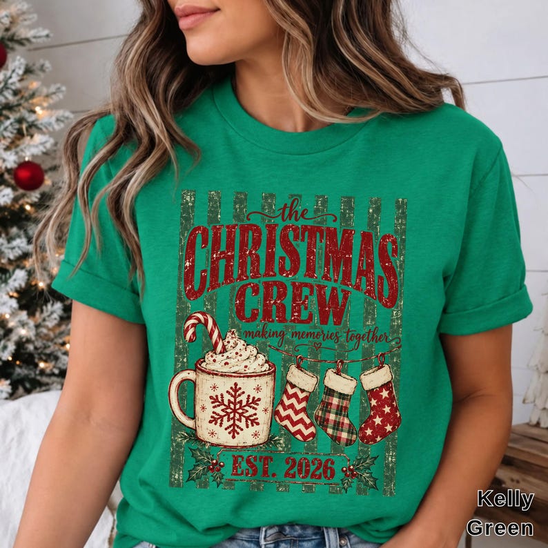 May include: A kelly green t-shirt featuring a graphic with the text "the CHRISTMAS CREW making memories together EST. 2026". The design includes a mug of hot cocoa with a candy cane, three hanging stockings, and holly leaves.