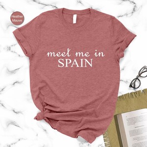 a red t-shirt with the text "Meet me in Spain" printed on it, along with a pair of glasses and a book on a marble surface.
