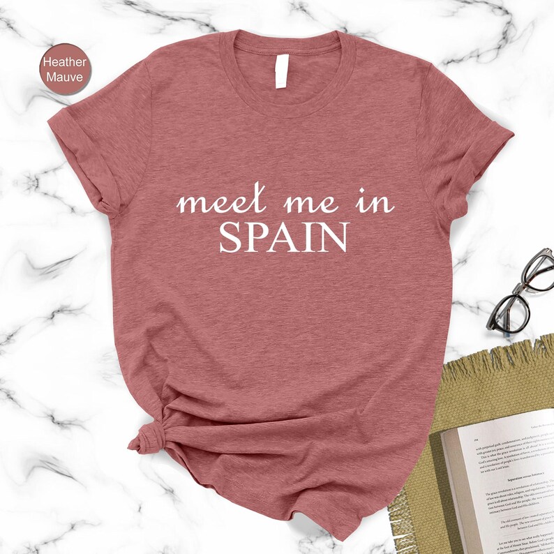a red t-shirt with the text "Meet me in Spain" printed on it, along with a pair of glasses and a book on a marble surface.
