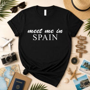 a black t-shirt with the text "Meet me in Spain" printed on it, surrounded by various travel-related items such as a passport, sunglasses, a hat, a camera, and a compass.