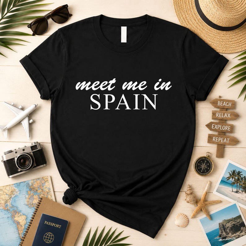 a black t-shirt with the text "Meet me in Spain" printed on it, surrounded by various travel-related items such as a passport, sunglasses, a hat, a camera, and a compass.