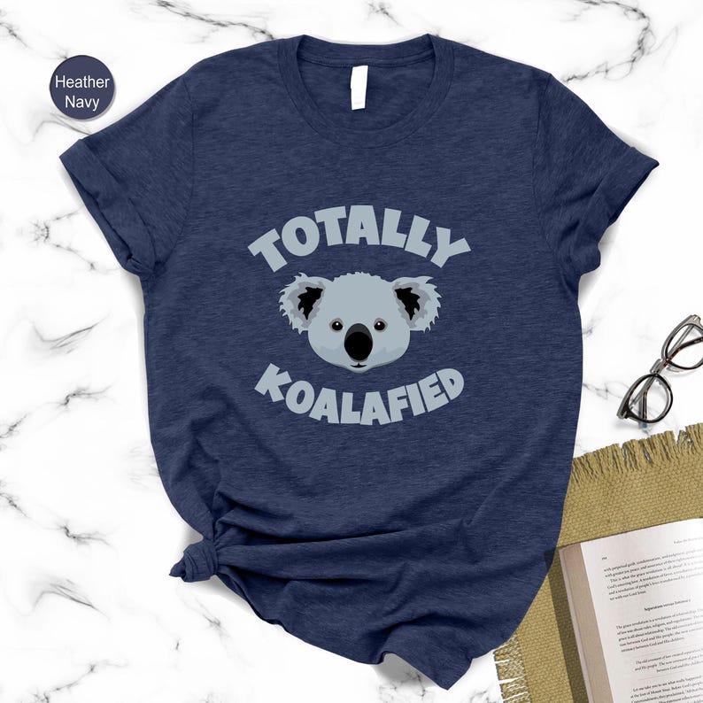 A navy blue t-shirt with a graphic of a koala and the text "Totally Koalified" printed on it, along with a pair of glasses and a book.