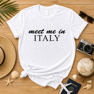 a white t-shirt with the text "Meet me in ITALY" printed on it, along with various travel-related items such as a hat, sunglasses, a camera, a globe, and seashells.