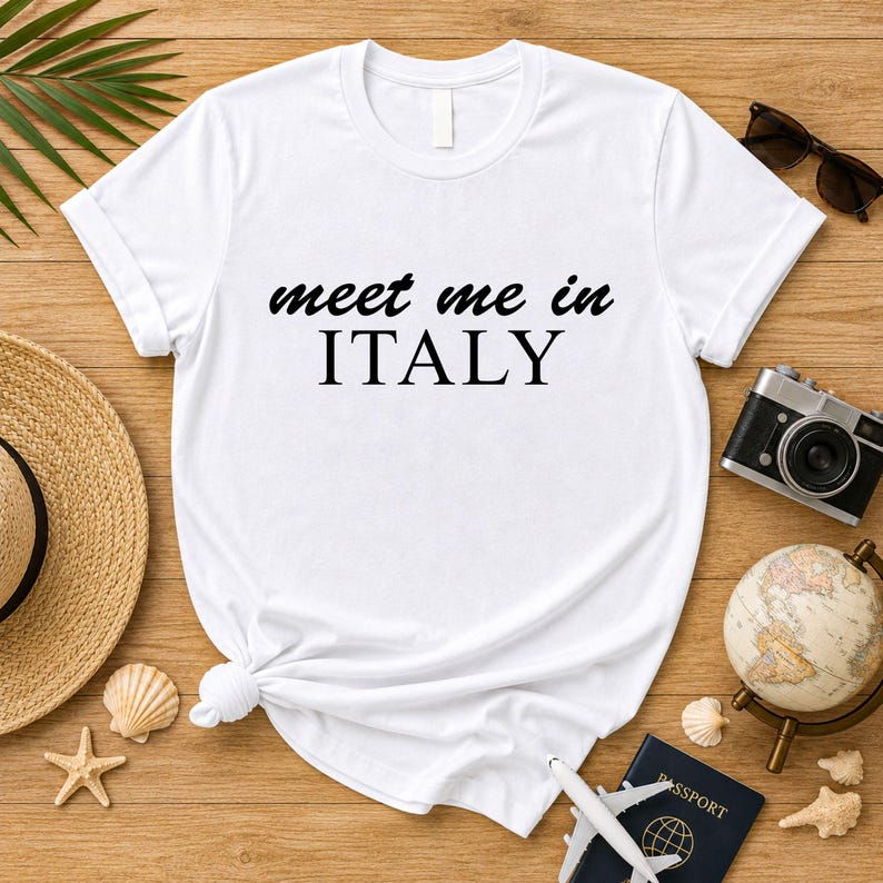 a white t-shirt with the text "Meet me in ITALY" printed on it, along with various travel-related items such as a hat, sunglasses, a camera, a globe, and seashells.