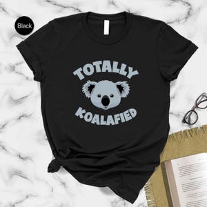A black t-shirt with a graphic of a koala and the text "Totally Koalified" printed on it, along with a pair of glasses and a book on a marble surface.