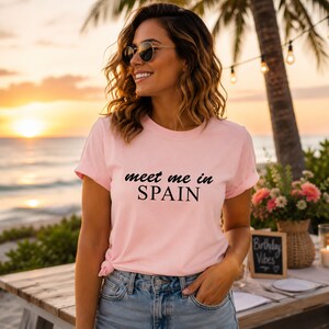 A woman wearing a pink t-shirt with the text "Meet me in Spain" stands on a beach at sunset, smiling and looking towards the camera.