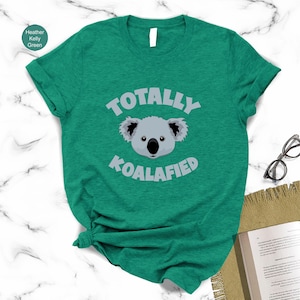 A green t-shirt with a cartoon koala and the text "Totally Koalified" printed on it, along with a pair of glasses and a book.