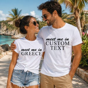 A man and a woman standing together, smiling and wearing matching t-shirts with the text "Meet me in Greece" printed on them.