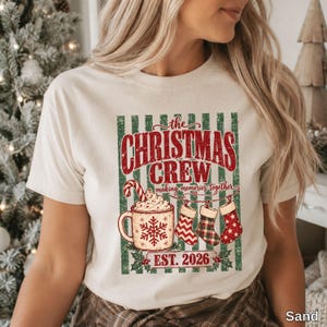 May include: A sand-colored t-shirt featuring a graphic with green vertical stripes and the text "the CHRISTMAS CREW making memories together EST. 2026". The design includes a mug with a snowflake, a candy cane, and three stockings.