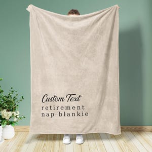 May include: A beige blanket with the words "Custom Text retirement nap blankie" in black script. The blanket is held up by a person, and the background is a solid teal color.