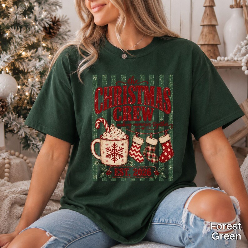 May include: A forest green t-shirt featuring a graphic with the text "the CHRISTMAS CREW" in red, a mug with a snowflake and candy cane, and three hanging stockings. Below the graphic, the text "EST. 2026" is printed in red.