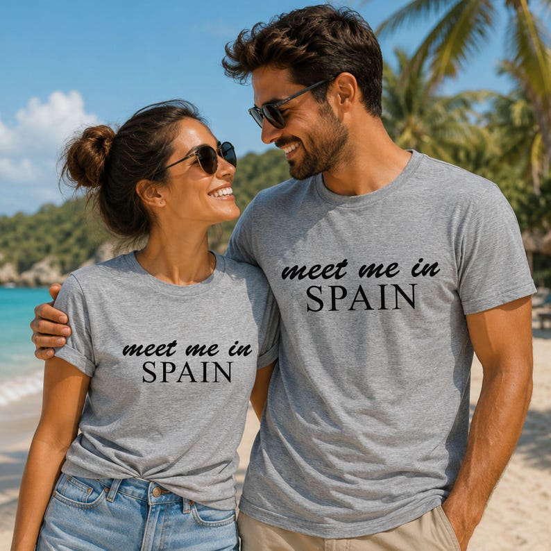 A man and a woman standing together on a beach, both wearing matching gray t-shirts with the text "Meet me in Spain" printed on them.