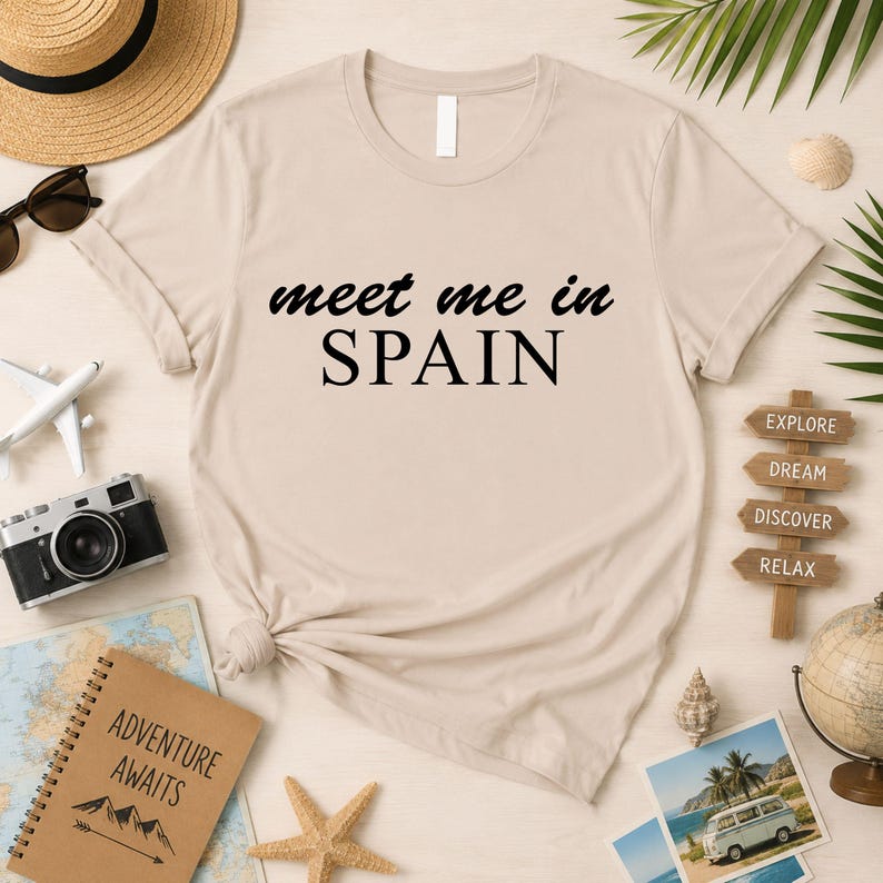 a beige t-shirt with the text "Meet me in Spain" printed on it, surrounded by various travel-related items such as a hat, sunglasses, a camera, a notebook, a globe, a starfish, a seashell, and a sign that says "EXPLORE DREAM DISCOVER RELAX".