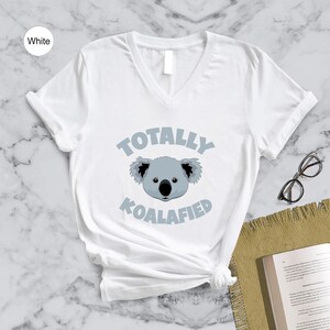 A white t-shirt with a graphic of a koala and the text "Totally Koalified" printed on it, along with a pair of glasses and a book on a marble surface.