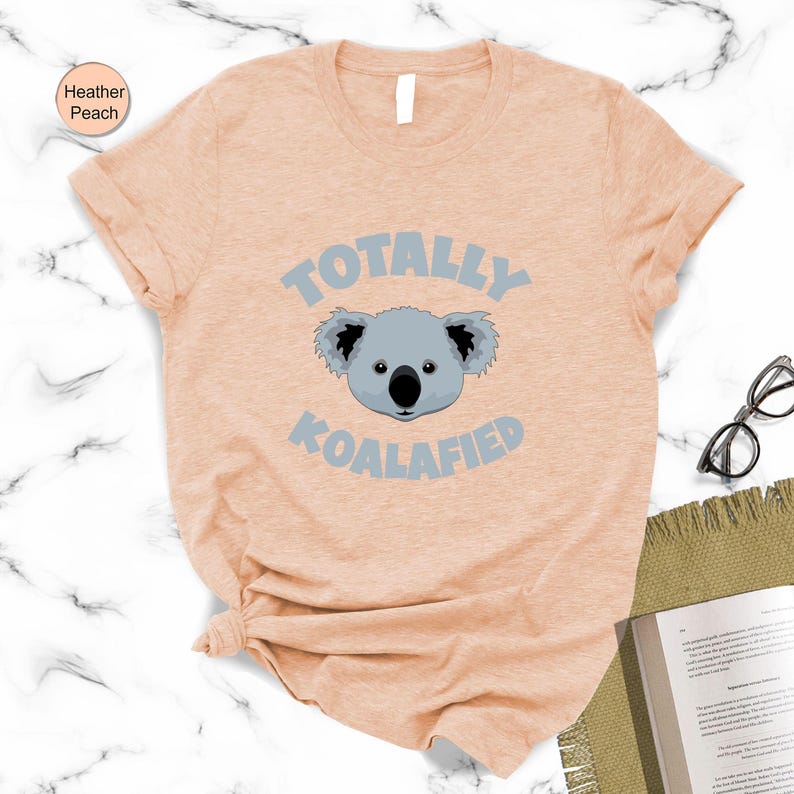 a light peach t-shirt with a graphic of a koala and the text "Totally Koalified" printed on it, along with a pair of glasses and a book.