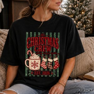 May include: A black short-sleeve t-shirt featuring a graphic with green vertical stripes, red text reading the CHRISTMAS CREW making memories together, a mug with a candy cane and snowflake, three hanging stockings, and EST. 2026.