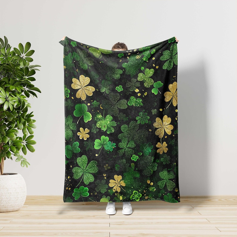 May include: A black blanket featuring a pattern of green and gold shamrocks. The shamrocks vary in size and are scattered across the blanket. Some shamrocks have a glittery appearance. The blanket is held up against a white wall.