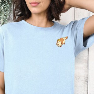 May include: A light blue t-shirt with a cartoon dog wearing a brown and white hat. The dog is looking to the left and has a brown nose and brown eyes.