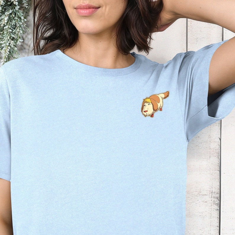 May include: A light blue t-shirt with a cartoon dog wearing a brown and white hat. The dog is looking to the left and has a brown nose and brown eyes.