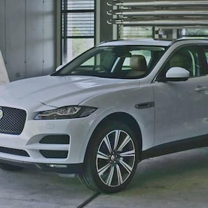 May include: A white Jaguar F-PACE SUV parked in a modern garage setting. The vehicle has a black grille, black and silver wheels, and tinted windows. The interior appears to be tan leather. The background includes trees and architectural elements.