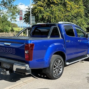 May include: A blue Isuzu D-Max pickup truck parked on a paved road. The truck has a black roll bar, silver side steps, and a black bed cover. The Isuzu logo is visible on the tailgate.