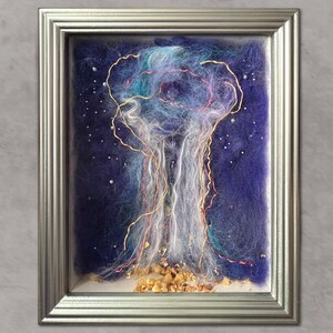 May include: A framed artwork featuring a felted abstract design. The piece has a dark blue background with white and light blue felted cloud-like shapes. Gold and pink wire accents and small gold flakes add detail. The artwork is encased in a silver frame.