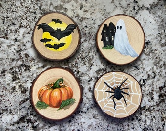 May include: Four circular wood slices featuring hand-painted Halloween designs. One shows black bats against a yellow moon, another a white ghost by a dark house, a third an orange pumpkin with green leaves, and the fourth a black spider on a web.