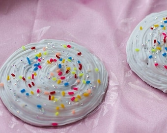 May include: Two white cupcake-shaped pasties with colorful sprinkles on a pink satin background.