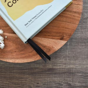 May include: A hardcover book with a light blue cover and a black bookmark rests on a wooden serving tray. The book is a New York Times bestseller. White flowers are in the upper left corner, and the background is a dark wood surface.