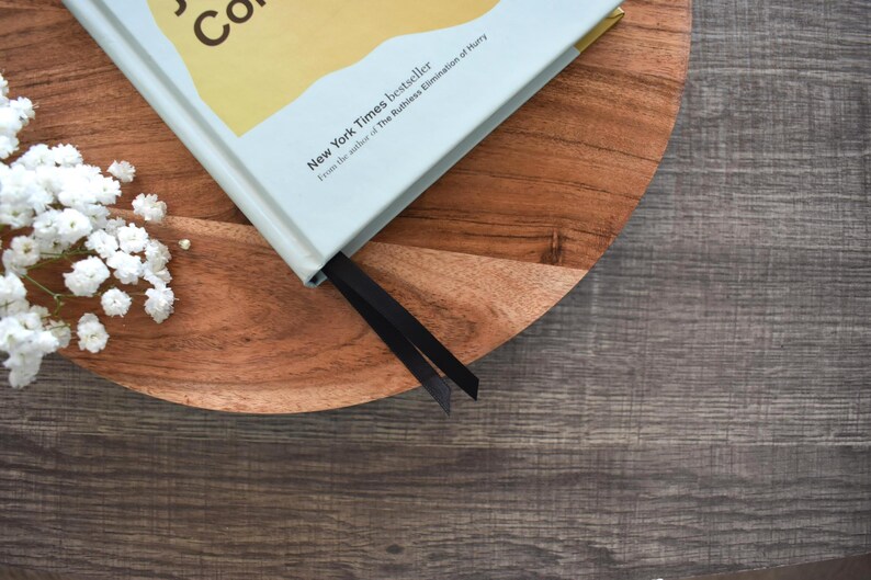 May include: A hardcover book with a light blue cover and a black bookmark rests on a wooden serving tray. The book is a New York Times bestseller. White flowers are in the upper left corner, and the background is a dark wood surface.