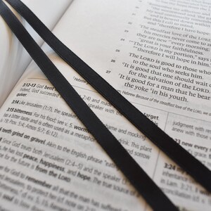 May include: Open book with black ribbon bookmarks. The pages are filled with text, and the ribbons are positioned diagonally across the open pages. The image has a soft, natural lighting.