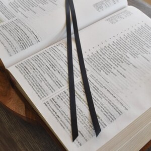 May include: An open book with black ribbon bookmarks. The pages are filled with text, and the book is resting on a wooden surface. White flowers are placed around the book.
