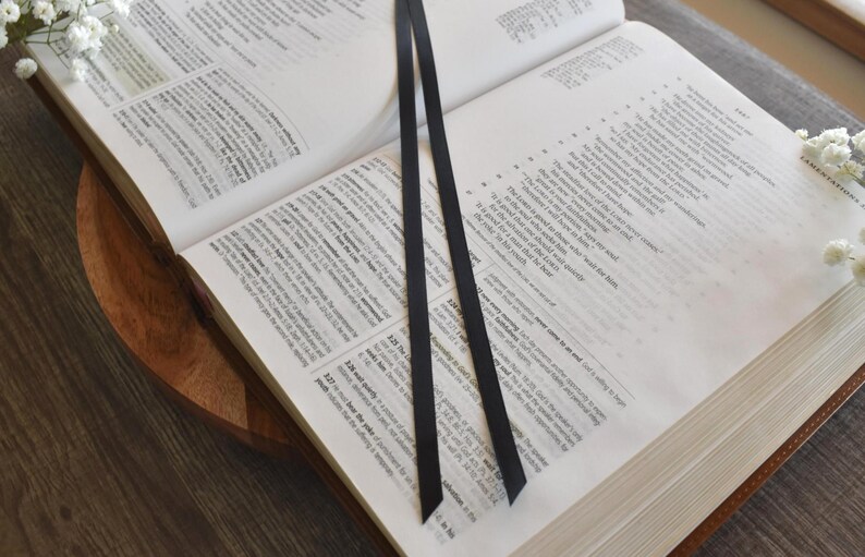 May include: An open book with black ribbon bookmarks. The pages are filled with text, and the book is resting on a wooden surface. White flowers are placed around the book.