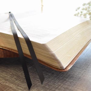 May include: An open book with gold-gilded edges and a brown leather cover. Two black ribbon bookmarks extend from the pages. The book rests on a wooden tray, with white flowers in the background.