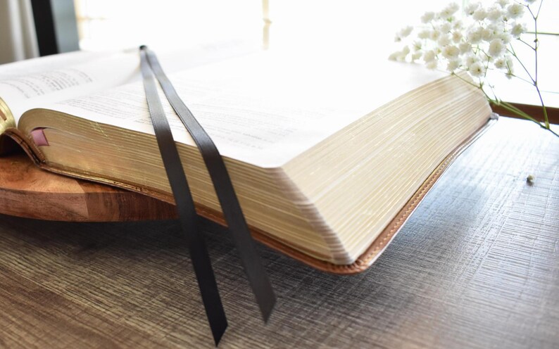 May include: An open book with gold-gilded edges and a brown leather cover. Two black ribbon bookmarks extend from the pages. The book rests on a wooden tray, with white flowers in the background.