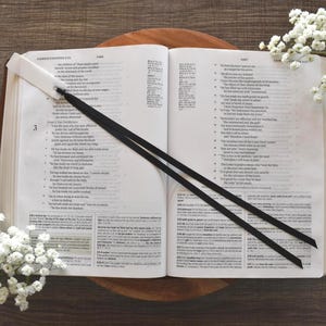 May include: An open book with text, featuring a black ribbon bookmark. The book rests on a wooden surface, with white flowers arranged around it. The pages are cream-colored, and the text is in black.