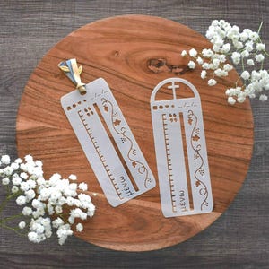 May include: Two white stencils with ruler markings and grape vine designs, one with a cross at the top. The stencils are on a wooden surface, with white flowers and a ribbon.